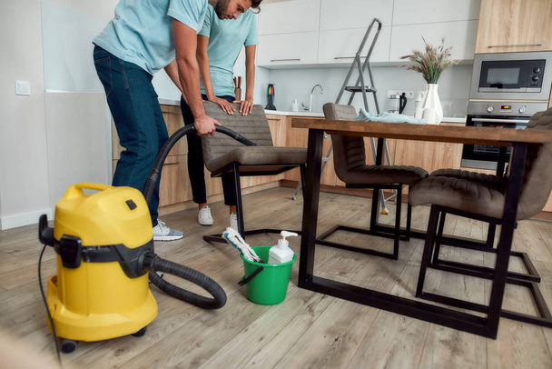 Обрезанная фотография двух молодых профессиональных уборщиков в униформе с помощью пылесоса при уборке кухонных стульев. Концепция услуг по уборке
 - Фото, изображение