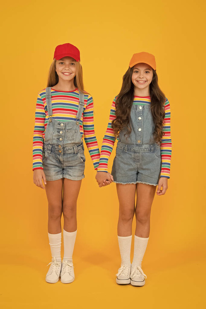 Friends stick together. Happy girls friends holding hands on yellow background. Adorable small friends enjoying friendship. Little friends smiling in fashionable overalls and stylish caps - Photo, image