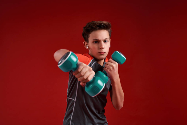 Power. A teenage boy engaged in sport, looking at camera while holding dumbbells. Isolated on red background. Training, fitness, active lifestyle concept. Horizontal shot - Valokuva, kuva