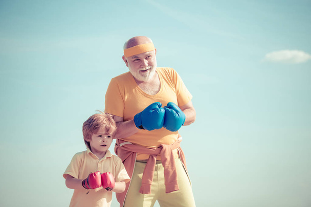 Elderly man hitting punching bag. Boxing. I love boxing. Portrait of a determined senior boxer with little boy child over blue sky background. - Фото, зображення