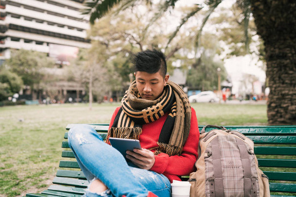 Portrait of young Asian man using his digital tablet while sitting in a bench outdoors. Technology concept. - Foto, Bild