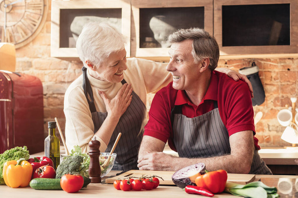 Coppia anziana che ride e si abbraccia mentre cucina il cibo in cucina insieme
 - Foto, immagini