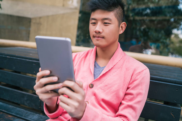 Portrait of young Asian man using his digital tablet while sitting in a bench outdoors. Technology concept. - Φωτογραφία, εικόνα