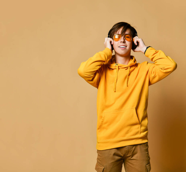 Young positive teen boy in yellow hoodie, sunglasses and headphones standing and smiling - Fotografie, Obrázek