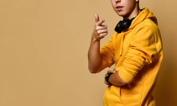 Young positive teen boy in yellow hoodie and headphones standing, smiling and looking at camera over yellow wall background - Photo, Image