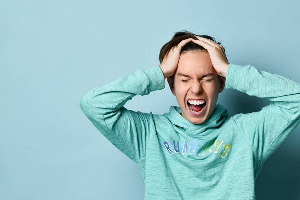 Retrato emocional de medio cuerpo de un adolescente caucásico con capucha azul. Tipo excitado gritando sosteniendo su cabeza en sus manos
 - Foto, Imagen
