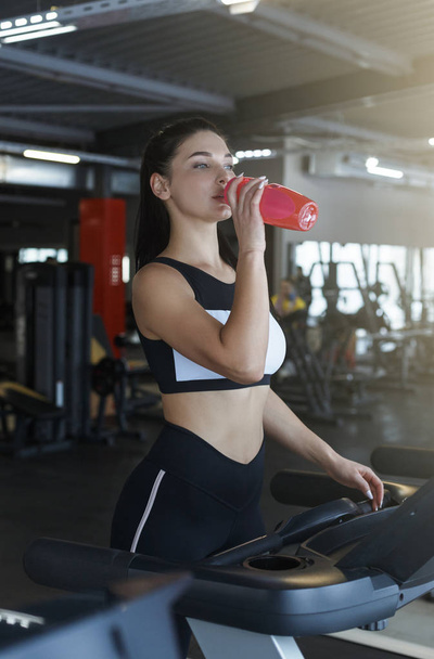 Mujer joven bebiendo agua o batido de proteínas mientras corre en la cinta de correr en el club deportivo
 - Foto, imagen