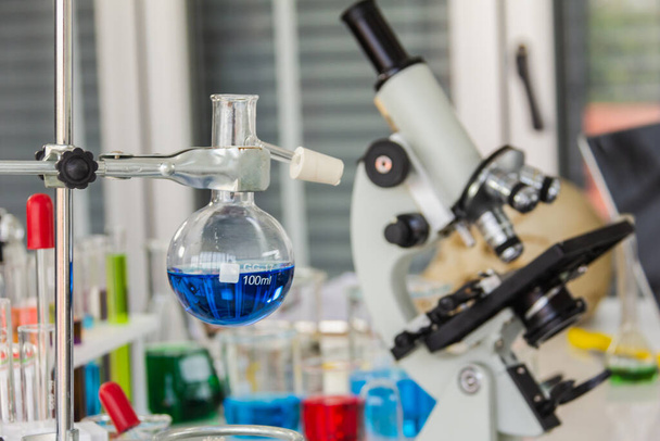 Óculos de destilação química com microscópios colocados em um laboratório com produtos químicos vermelhos, laranja, azuis no copo e tubos colocados na mesa de operação
 - Foto, Imagem