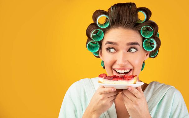 Hungry Girl With Curlers Eating Cake Posing On Yellow Background - 写真・画像