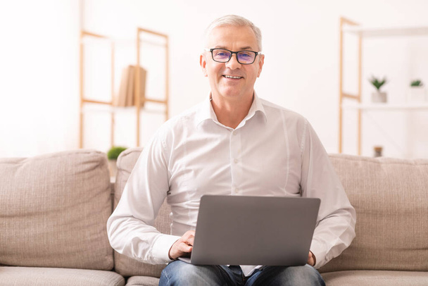 Uomo anziano che utilizza il computer portatile e seduto sul divano a casa
 - Foto, immagini