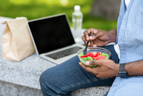 Невідомий чоловік їсть овочевий салат та використовує ноутбук на відкритому повітрі
 - Фото, зображення