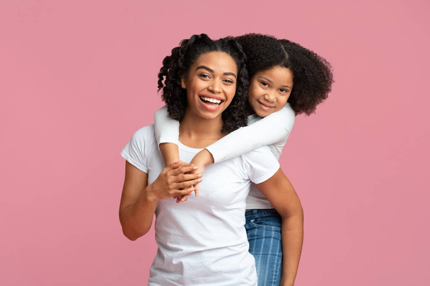 Маленькая черная девочка обнимает свою мать сзади на розовом фоне
 - Фото, изображение