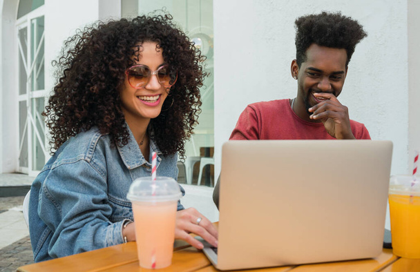 Portret dwóch afro przyjaciół korzystających z laptopa podczas picia świeżego soku owocowego na świeżym powietrzu w stołówce. - Zdjęcie, obraz