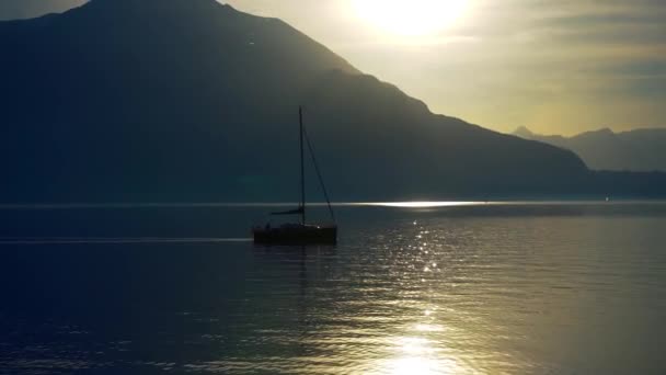 セーリングヨットは、イタリアのコモ湖の海岸沖で係留され、高山の山の地平線の上に日没前に係留されました。シネマティック・フレーム。ロマンチックな雰囲気。静かで平和的なシーン。帆船 - 映像、動画