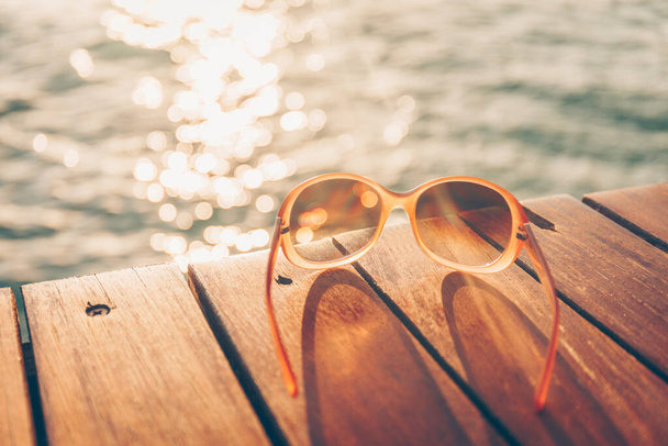 Okulary przeciwsłoneczne na drewnianym molo podczas zachodu słońca. Luksusowy ośrodek wypoczynkowy. Koncepcja wakacyjnego wyjazdu - Zdjęcie, obraz