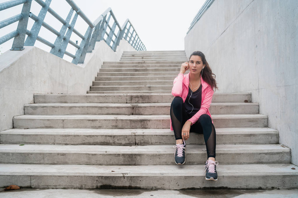 Portrait of an athletic woman listening to music on a break from training while sitting on stairs. Sport and health lifestyle concept. - Photo, Image