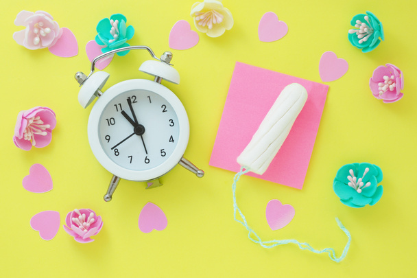 un pequeño despertador blanco, un tampón, corazones rosados y flores artificiales multicolores. el concepto de menstruación, ovulación, anticoncepción
 - Foto, imagen