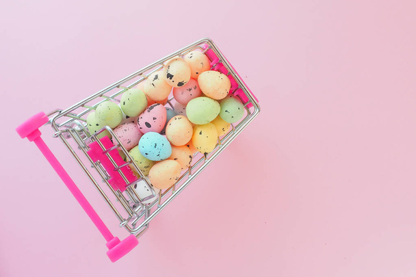 πολλά πολύχρωμα μικροσκοπικά τεχνητά αυγά σε ένα μικροσκοπικό καλάθι τροφίμων σε ροζ φόντο. Θέα από ψηλά. Έννοια πώλησης Πάσχα - Φωτογραφία, εικόνα