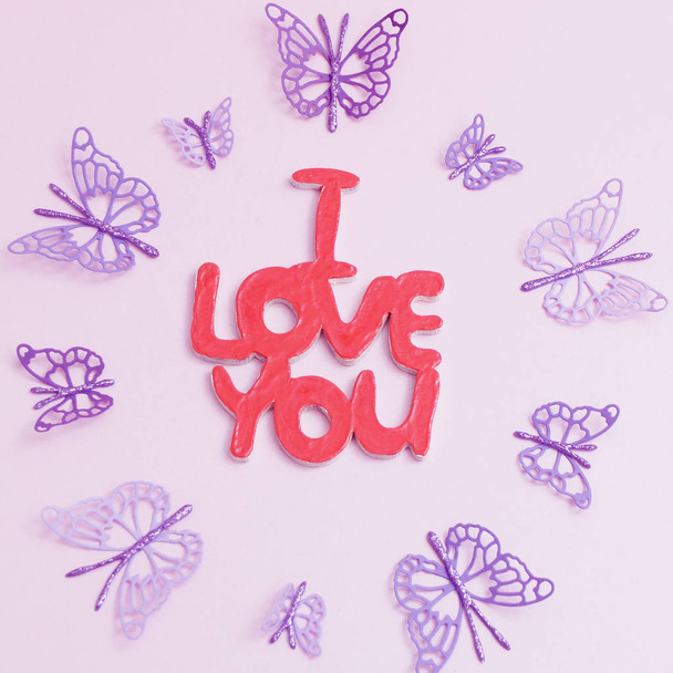 Inscripción de madera "I love you" sobre un fondo rosa y mariposas de papel morado alrededor
 - Foto, Imagen