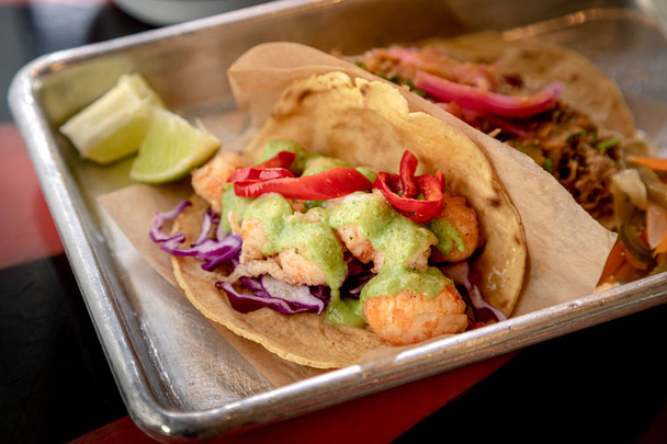 Close up of shrimp tacos in flour tortillas with verde sauce sitting on metal platter lined with brown paper liner - Photo, Image