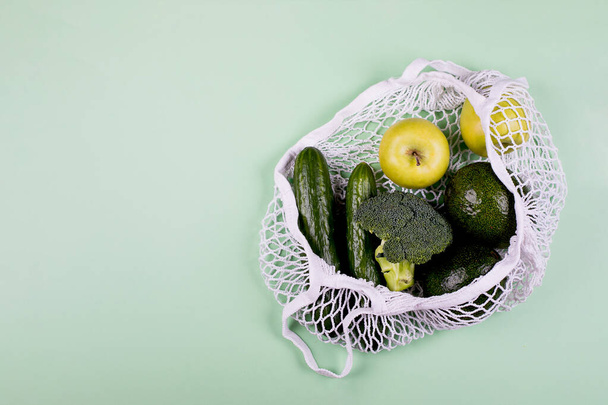 緑のテーブルの背景に緑の野菜や黄色の果物とメッシュショッピングバッグ。トップビュー、フラットレイアウト、コピースペース。廃棄物ゼロ、環境に優しいコンセプト. - 写真・画像