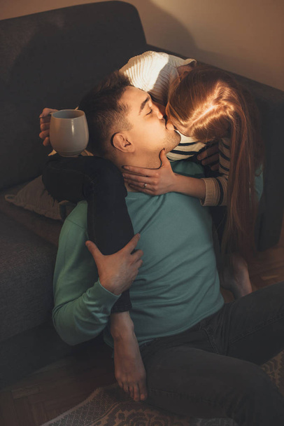 Charming caucasian girl with red hair is kissing her lover while holding a cup of tea during a sunny morning - Photo, Image