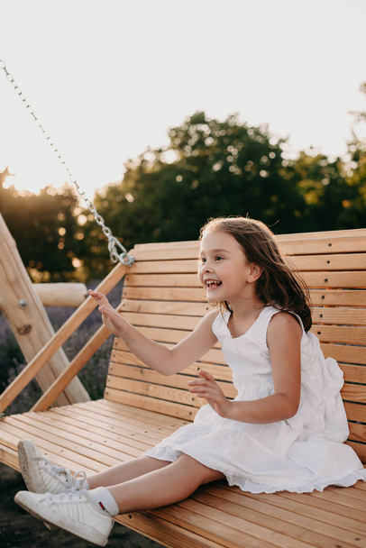 Cheerful caucasian girl dressed in a white dress is sitting in a swing having fun in a summer evening - Фото, зображення