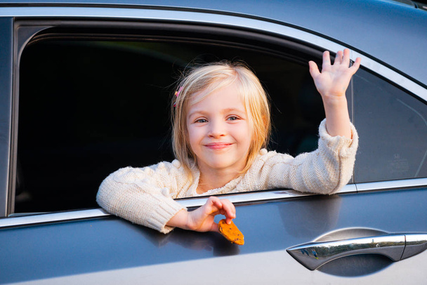 Pieni tyttö työntää päänsä ulos auton ikkunasta. Automatka tai matka käsite. Onnelliset lapset kulkevat autolla. Söpö lapsi nojaa ulos pakettiauton ikkunasta, hymyilee ja vilkuttaa
. - Valokuva, kuva