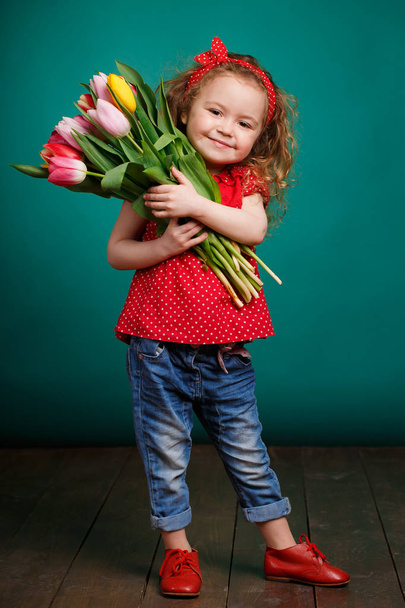 Spring portrait of a smiling little girl with red curly hair holding a bouquet of multi-colored tulips as a gift, isolated on a light green background in the Studio.A cute little girl with a bouquet of tulips, in the spring in the studio. - Fotografie, Obrázek