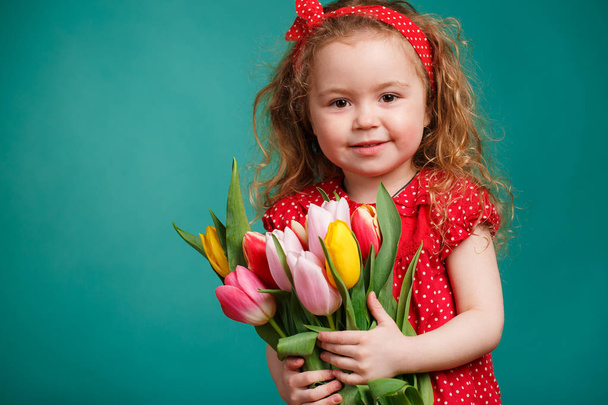 Spring portrait of a smiling little girl with red curly hair holding a bouquet of multi-colored tulips as a gift, isolated on a light green background in the Studio.A cute little girl with a bouquet of tulips, in the spring in the studio. - Foto, imagen