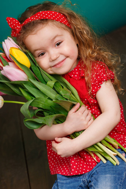 Spring portrait of a smiling little girl with red curly hair holding a bouquet of multi-colored tulips as a gift, isolated on a light green background in the Studio.A cute little girl with a bouquet of tulips, in the spring in the studio. - 写真・画像