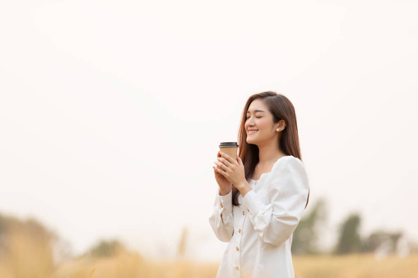 Young woman holding takeaway coffee cup standing outdoors at barley field land in the morning. - Photo, Image