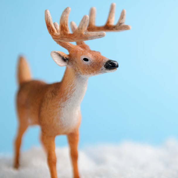 Toy deer on blue background is standing on artificial snow close-up. Background for childrens or winter advertising banner. - Fotoğraf, Görsel