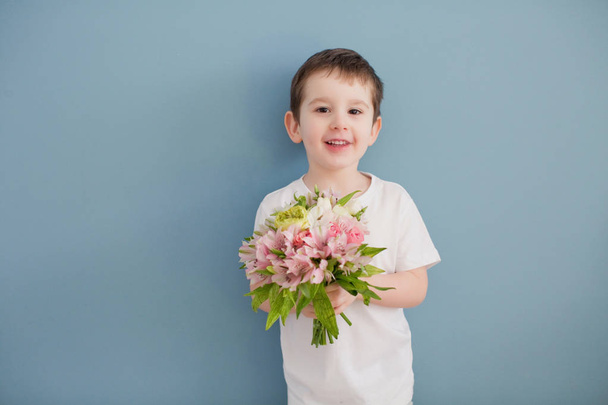 Petit garçon en t-shirt blanc tient bouquet de fleurs roses. Enfant drôle sur le fond du mur bleu
 - Photo, image