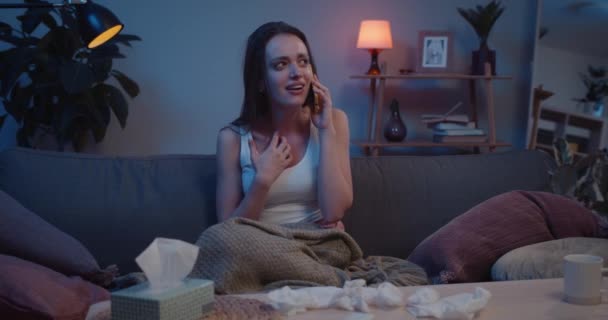 Young sad woman talking on phone and covering face with hand after brokeup with boyfriend. Front view of unhappy girl sitting on sofa, crying and having conversation. Home background. - Materiał filmowy, wideo