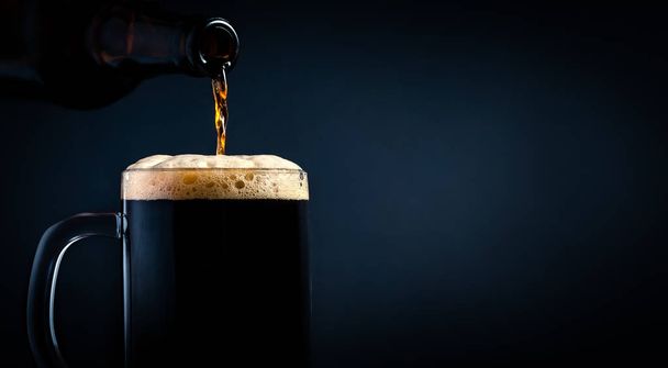 Ein Becher Schwarzbier, der in Großaufnahme aus einer Flasche auf dunklem Hintergrund ergießt. Archivbild. - Foto, Bild