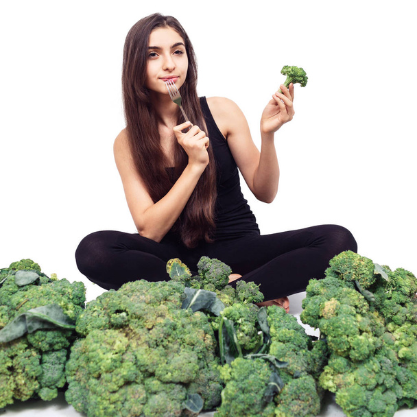 Genç ve güzel bir kız büyük bir brokoli yığınının içinde oturuyor ve dudağına çatal dayıyor. Sağlıklı beslenme konsepti. Saç ve deri sağlığı. beyaz üzerine izole. - Fotoğraf, Görsel