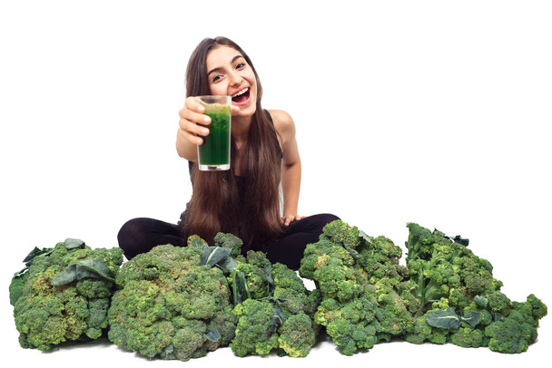 Neşeli vejetaryen kız brokolinin arasında oturuyor, elinde yeşil bir meyve püresi tutuyor ve onu size sunuyor. beyaz üzerine izole. - Fotoğraf, Görsel