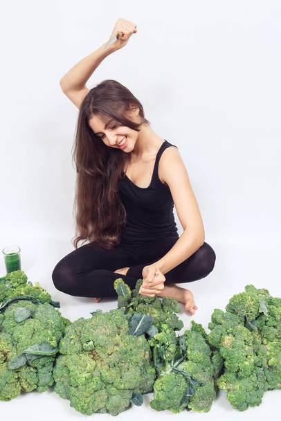 young beautiful girl sits in big heap of broccoli and pricks it on fork. Healthy eating concept. hair and skin health - Fotografie, Obrázek