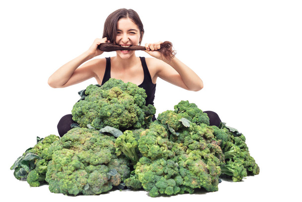 Genç ve güzel bir kız büyük bir brokoli yığınına oturup saçlarını ısırıyor. Sağlıklı beslenme konsepti. Saç ve deri sağlığı. beyaz üzerine izole. - Fotoğraf, Görsel