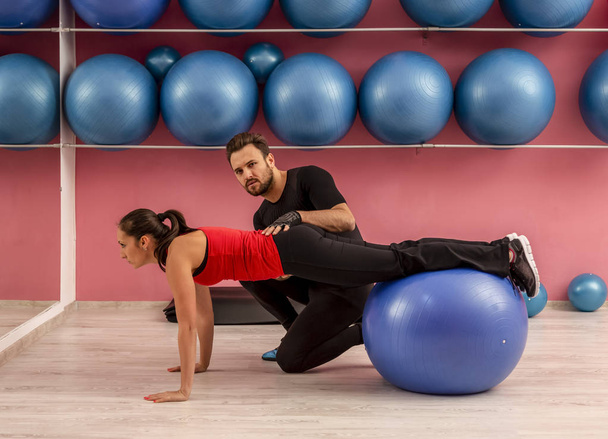 Young woman helped by her coach is doing exercises with a fitness ball  in a gym. - Φωτογραφία, εικόνα