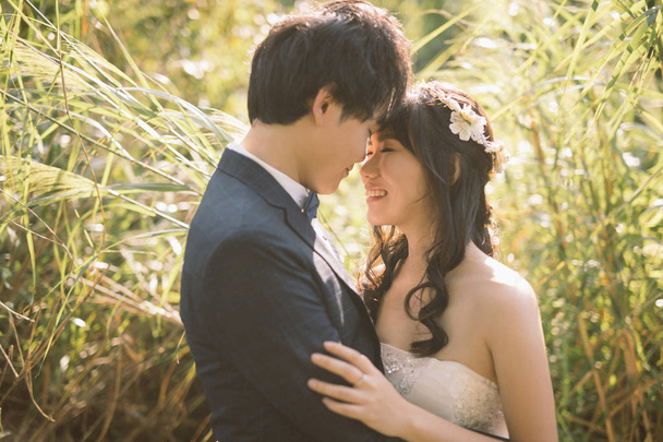 happy bride and groom embracing and looking each other while posing among meadow at sunny day , wedding concept   - Photo, Image