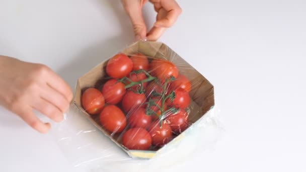 Frau verwendet Lebensmittelfolie zur Lagerung von Lebensmitteln auf einem weißen Tisch. Rolle aus transparentem Polyethylen für Lebensmittelverpackungen. Kirschtomaten in Einweg-Plastikverpackung. Zeitlupenvideo. Nahaufnahme - Filmmaterial, Video