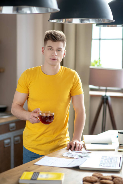 Nuori vaalea mies keltaisessa t-paidassa seisoo pöydän lähellä
 - Valokuva, kuva