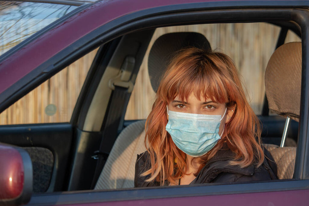 女の子は車の中で医療マスクを着ている。コロナウイルスから保護するために、コビト-19と書かれたマスクを身に着けている女性。ウイルスのパンデミックの間にマスクで車を運転。マスクは毎日の活動に置く必要があります。. - 写真・画像