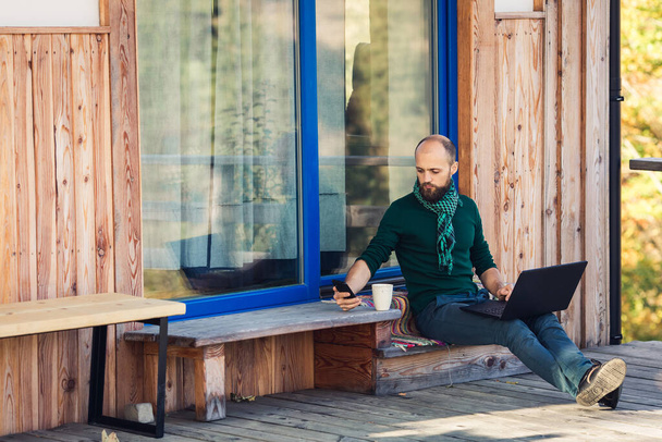 bearded freelancer works on laptop and talks on phone, sitting on steps of private house - Photo, Image