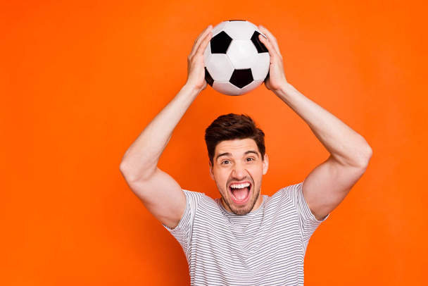 Photo of attractive crazy guy hold football leather ball hands above head supporting team watch television wear striped t-shirt isolated bright orange color background - Foto, immagini