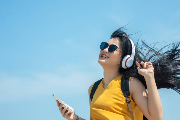 Mulher asiática feliz com mochila usando fone de ouvido na praia, conceito de estilo de vida
. - Foto, Imagem