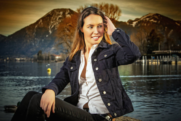 Жінка сидить на березі озера з джинсовою курткою і джинсами
 - Фото, зображення