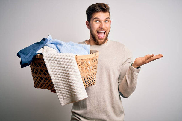 Young blond man with beard and blue eyes doing chores holding wicker basket with clothes very happy and excited, winner expression celebrating victory screaming with big smile and raised hands - Photo, Image